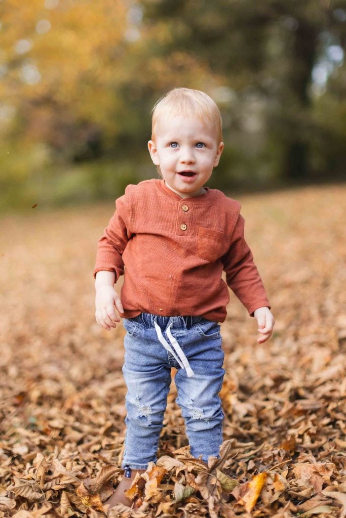 A toddler with blonde hair stands in a field of fallen leaves, wearing a rustcolored longsleeve shirt and blue jeans. The scene evokes the charm of a Bowie portrait with trees displaying autumnal foliage in the blurred background.