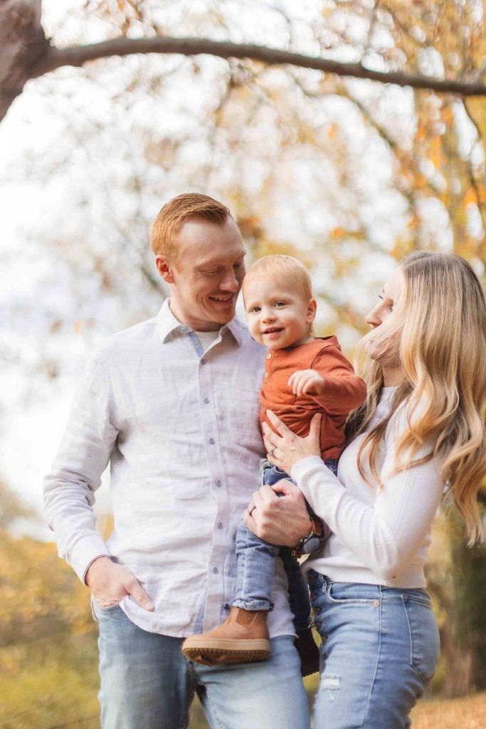 A man and woman standing outdoors in Bowie, Maryland, smiling and holding a young child. The man and child have red hair, and the woman has long blonde hair. Trees with fall foliage are in the background, adding to the picturesque scene near Belair Mansion.