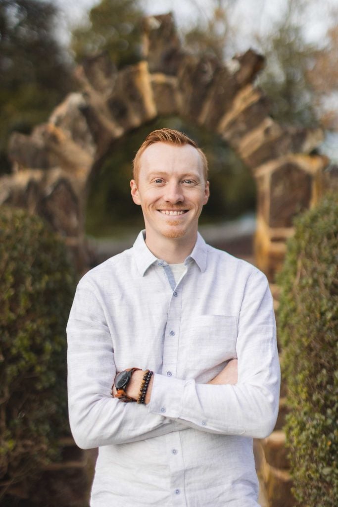 A redhaired man wearing a white shirt stands with folded arms in front of a rustic stone archway at Belair Mansion in Bowie, smiling. Bushes flank the archway, adding a charming touch to the historic family estate.