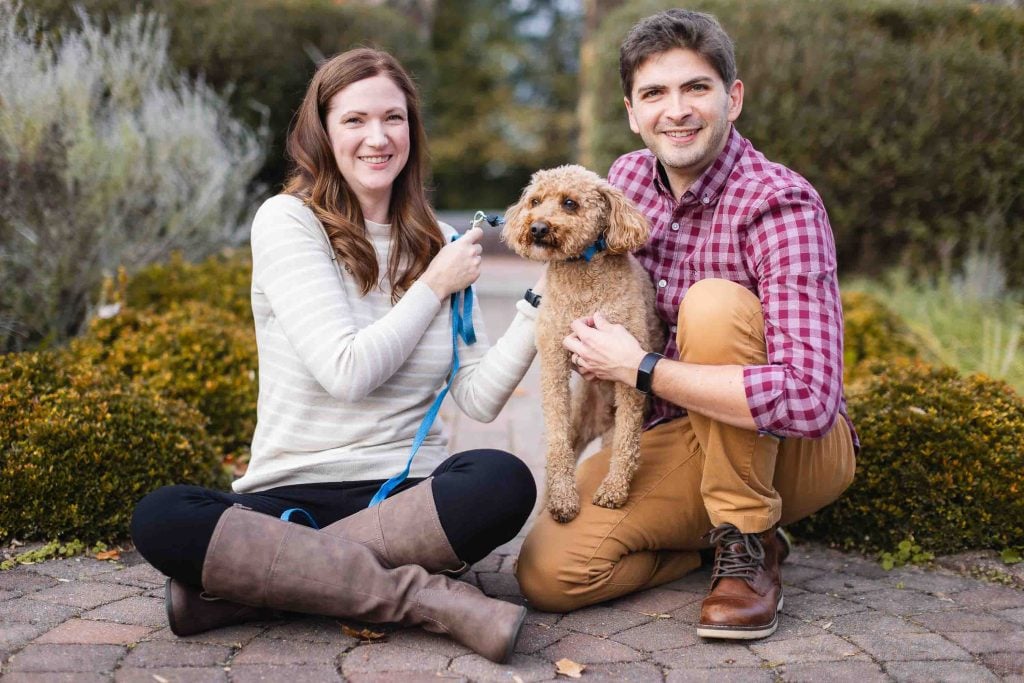 A man and a woman are outdoors in Maryland, both smiling while holding a small dog. The woman is sitting on the ground holding a leash, and the man is crouching down next to the dog, capturing the perfect moment during their minisession.