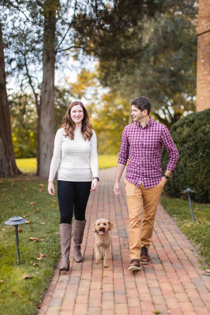 A man and woman walk down a brick pathway in Maryland with a small dog between them. Trees and bushes are in the background. They are smiling, casually dressed, and enjoying their minisession together.