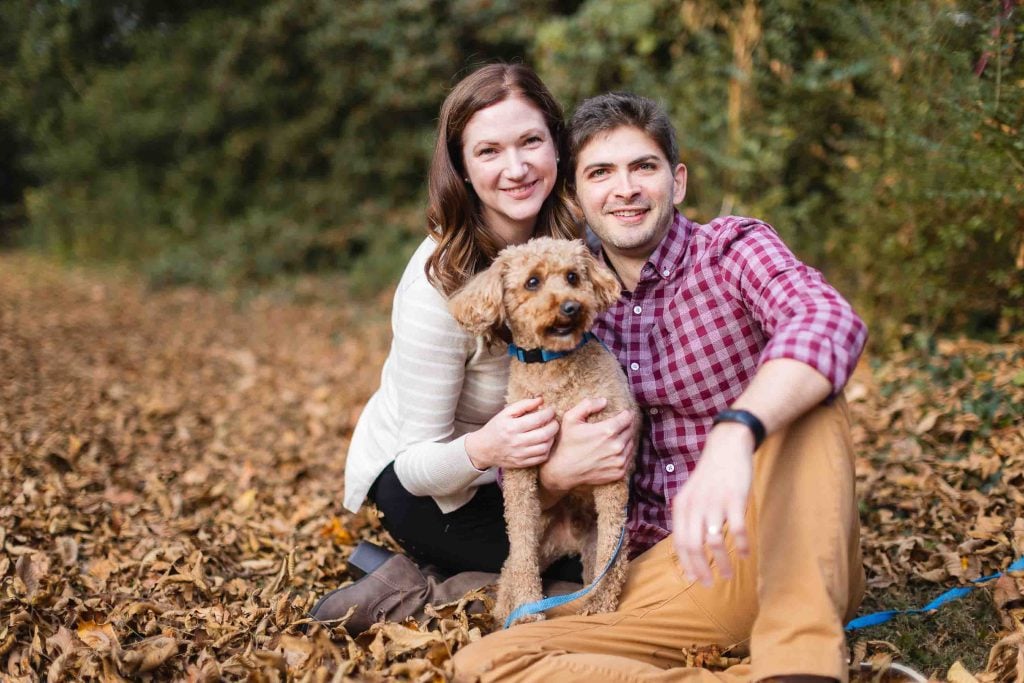 A woman and man sit on the ground among fallen leaves, smiling at the camera in a beautiful family portrait, while holding a small dog.