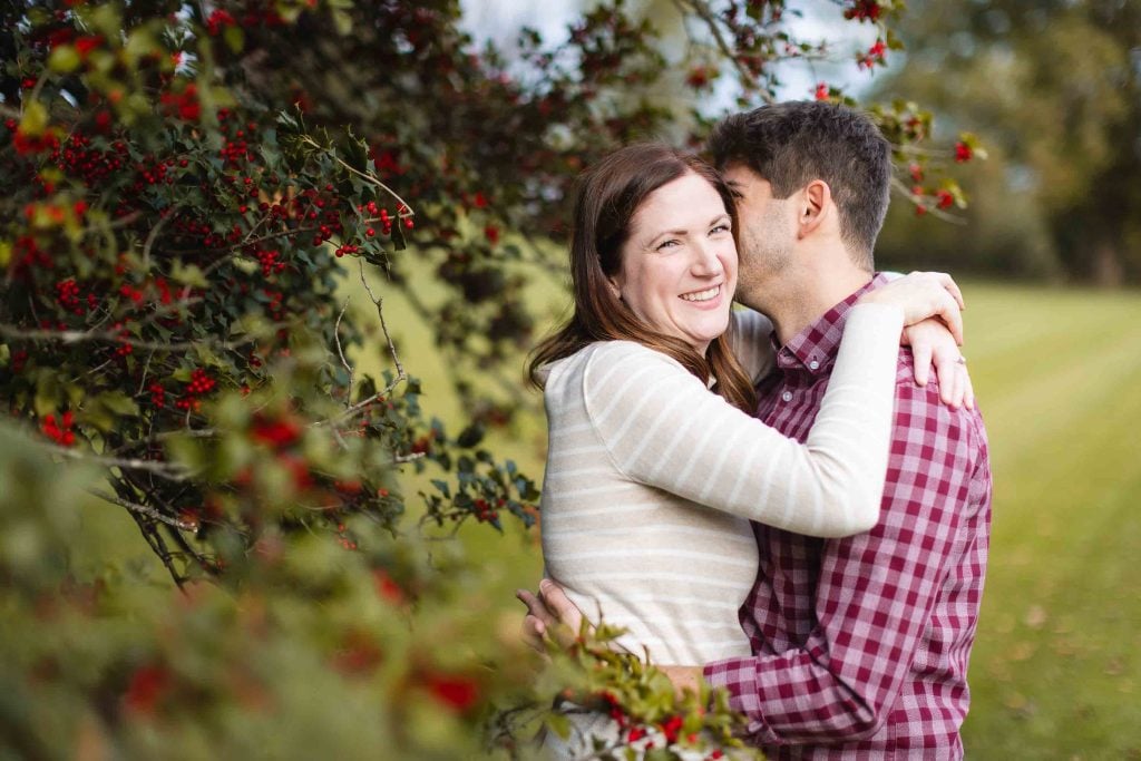 A couple is embracing outdoors near a berryladen bush. The woman is smiling while the man, dressed in a red checkered shirt, has his face partially hidden. This tender moment was captured during a minisession in Bowie, preserving a beautiful memory for their family.