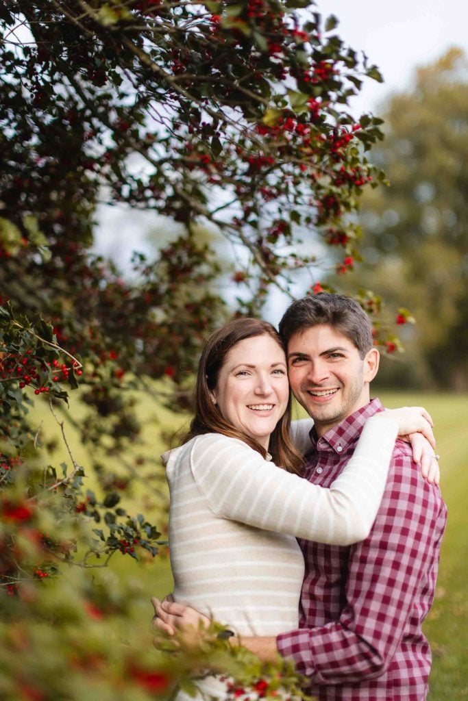 A couple, both smiling, stands outdoors embracing near a bush with red berries at Belair Mansion in Bowie, Maryland.