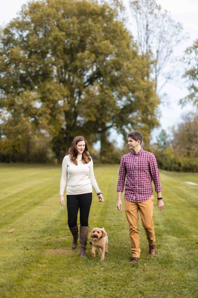 A man and woman walk a small dog on a grassy path with trees in the background, capturing a serene moment perfect for a minisession at Bowie’s historic Belair Mansion.