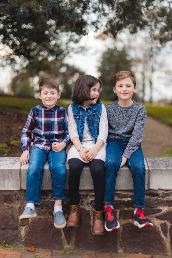 Three children sit on a stone wall in a park in Maryland, surrounded by greenery. Casually dressed and smiling, it’s a charming family portrait.