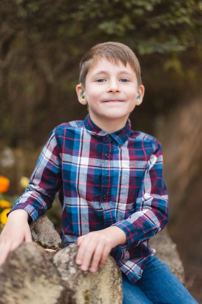 A young boy wearing hearing aids and a plaid shirt smiles while leaning against a stone structure at the historic Belair Mansion in Maryland.