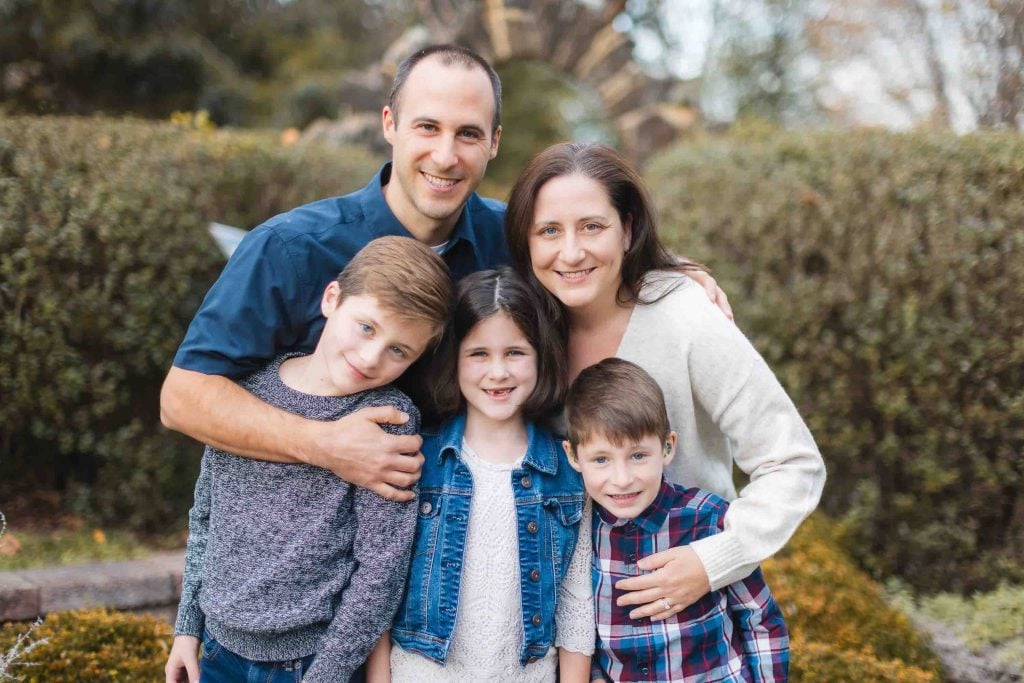 A group photo of a family during a minisession in Maryland, featuring two adults and three children standing close together and smiling at the camera, with greenery in the background.