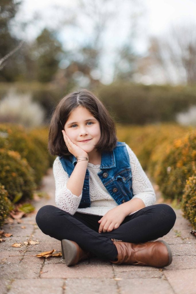 A girl with dark hair sits crosslegged on a paved path, resting her head on her hand. She is wearing a denim vest over a white sweater, reminiscent of Bowie's relaxed style, and there are hedges in the background. It's the perfect scene for a portrait minisession.