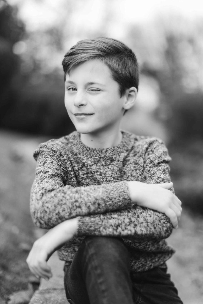 Black and white image of a young boy with short hair sitting outdoors in Bowie, Maryland, crossing his arms and winking at the camera. He's wearing a knitted sweater. The background is blurred, focusing on the boy. A precious family moment captured perfectly.