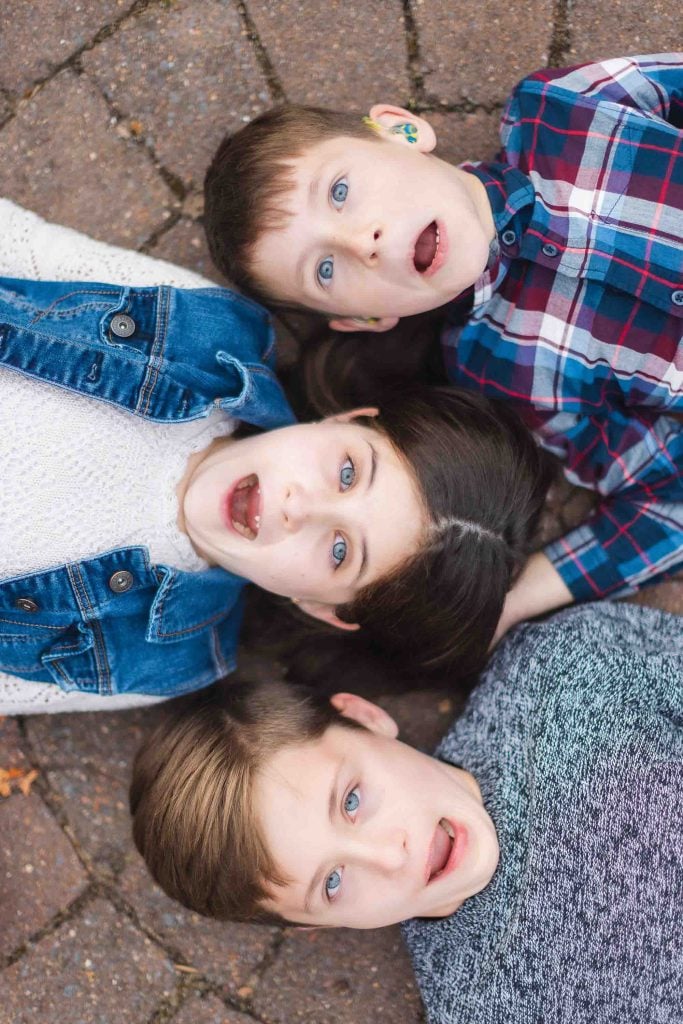 Three children lying on a brick surface, dressed in casual clothing, with surprised expressions on their faces. They are positioned closely together, heads touching in a triangular arrangement, reminiscent of a Bowie album cover—a snapshot capturing the spirit of family at Belair Mansion.