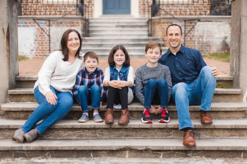 A family of five sits on the outdoor steps of the Belair Mansion in Maryland, smiling. The group includes two adults and three children; the adults are on either side, with the children in the center.