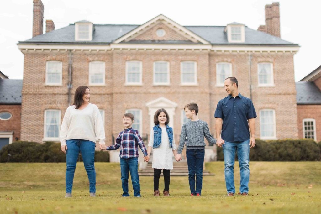 A family of five, consisting of a mother, father, and three children, stand holding hands on a lawn in front of the historic Belair Mansion in Bowie, Maryland.