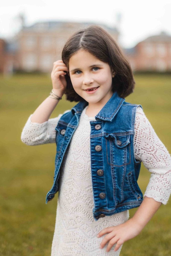 Young girl with dark hair, wearing a white dress and denim vest, stands outdoors on a grassy field near Belair Mansion, smiling slightly and touching her hair. Blurred buildings are visible in the background of the minisession portrait.