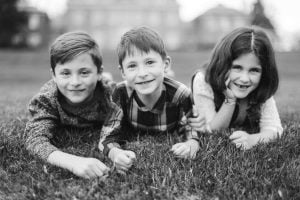 Three children lie on the grass, smiling at the camera. They appear to be outdoors, with a blurry building and trees visible in the background. The image feels like a timeless family portrait, possibly taken near Belair Mansion. The photograph is in black and white.