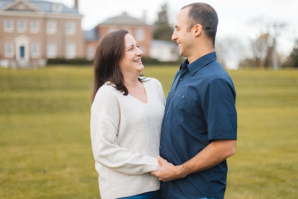 A couple stands outdoors on a grassy area in Maryland, smiling and looking at each other, with the grand Belair Mansion in the background.