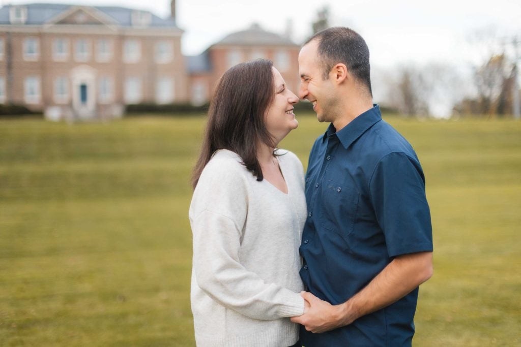 A couple stands close, smiling and holding hands, in an outdoor setting with green grass and a large building in the background of their Bowie, Maryland home.