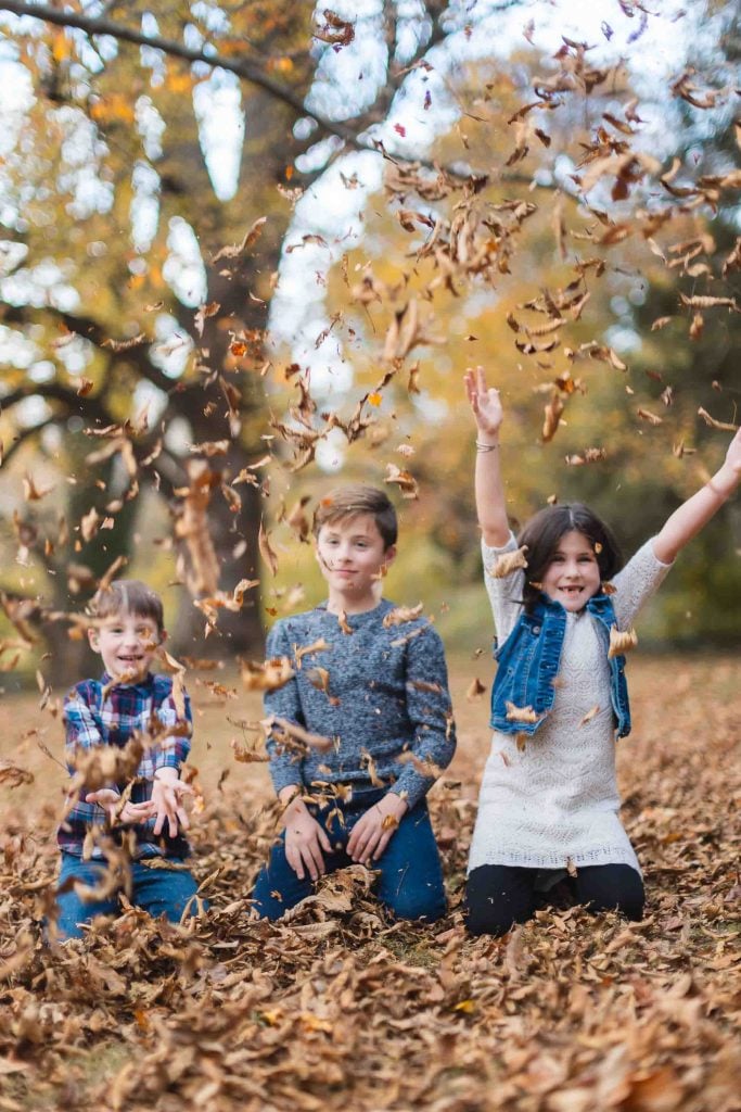 Three children play with fallen leaves in an autumn forest near Belair Mansion in Bowie, throwing leaves into the air and smiling, creating cherished family memories.