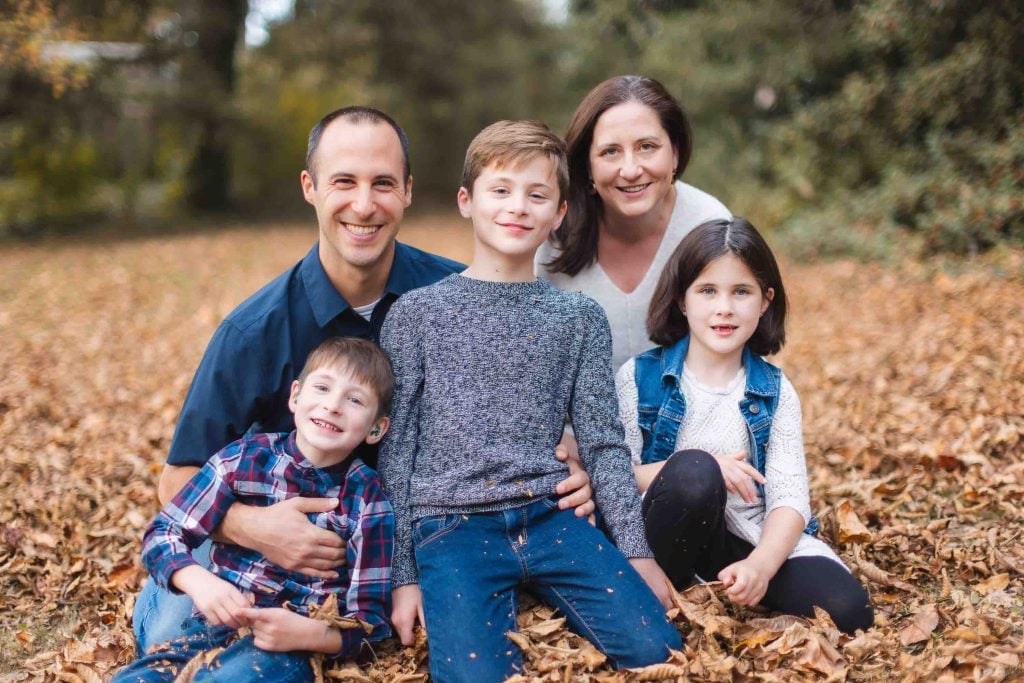 A family of five, including two adults and three children, sits on a ground covered in autumn leaves in Bowie, Maryland, smiling and posing for an outdoor photo.