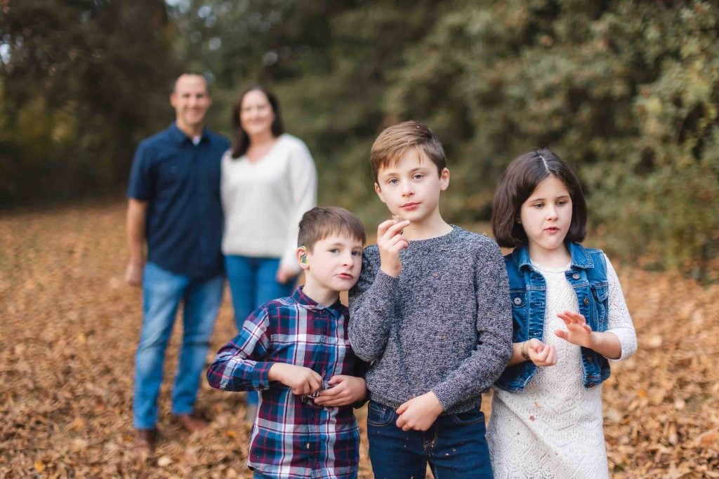 Three children stand together in a wooded area with fallen leaves during a family minisession, while two adults stand blurred in the background.