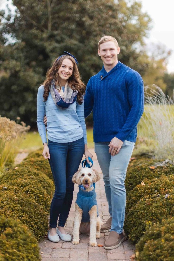 A woman and a man, both wearing blue tops, stand on a garden path at Belair Mansion with their dog. The dog is also dressed in a blue sweater. Shrubs and trees are in the background, setting the perfect scene for a minisession family photo.