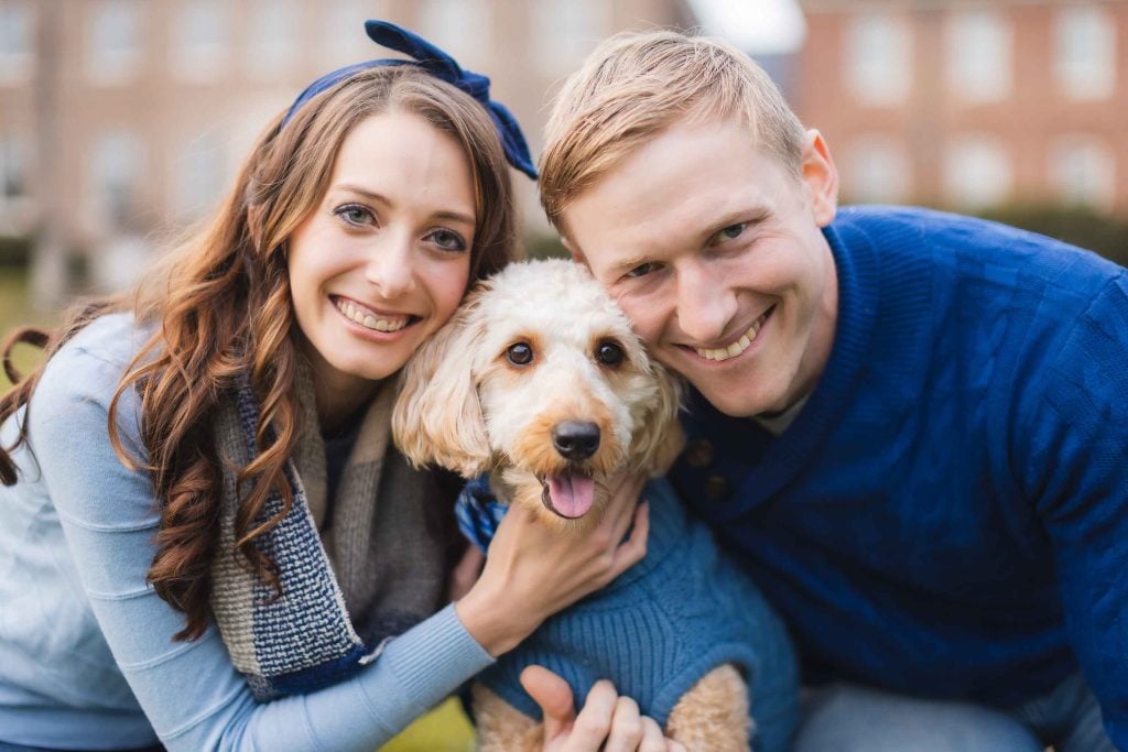 A man and woman, both smiling, pose closely with a small, happy dog wearing a blue sweater. They are outside in front of a brick building that resembles the historic Belair Mansion.