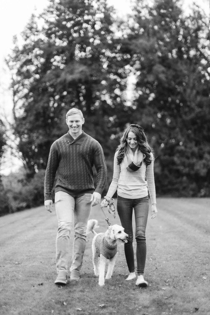 A man and a woman walk a dog on a leash in a grassy area with tall trees in the background. The image, capturing this serene family minisession, is rendered in black and white.