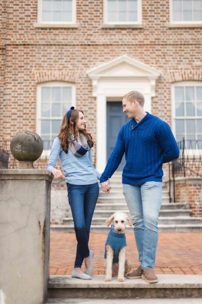 A family stands holding hands on the steps of a brick house, with a dog in a blue sweater sitting between them, capturing the perfect moment for their Maryland minisession.