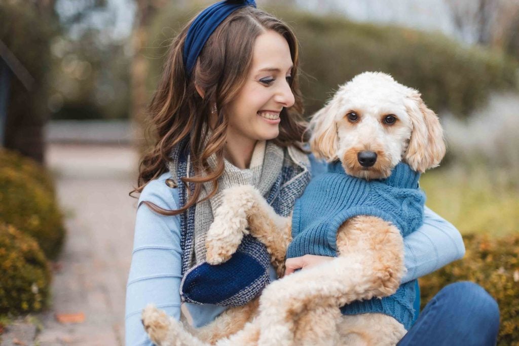 A woman in a blue sweater and headband is sitting outdoors, holding a creamcolored dog wearing a matching blue sweater. Both seem content during their minisession, with the blurred greenery of Belair Mansion in the background.