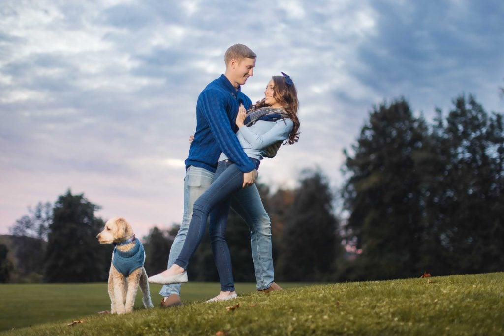 A couple dressed in blue are on a grassy hill in Maryland, with the man dipping the woman backward in a playful pose while a dog in a blue sweater stands nearby. This charming portrait captures their joy and the serene landscape.