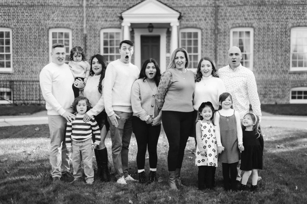 Black and white portrait of a large, smiling family posing in front of a brick building with windows and an arched entrance at Belair Mansion.