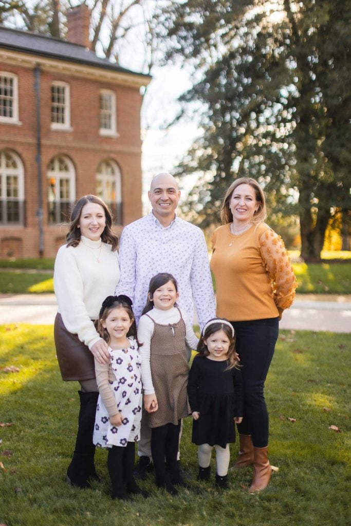 A family of six, including four adults and two children, stands on the lawn in front of the Belair Mansion on a sunny day. The adults are smiling, and the children stand in front dressed in casual clothing, capturing a timeless portrait of joy and togetherness.