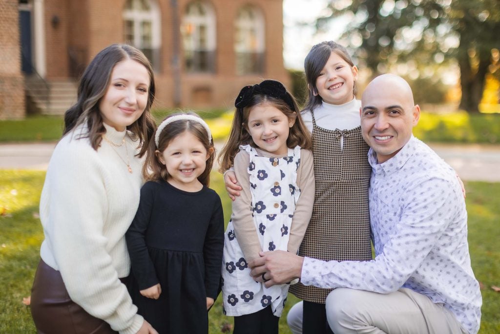 A portrait of a man and woman with three young girls smiling while posing for a photo outdoors in front of the Belair Mansion.