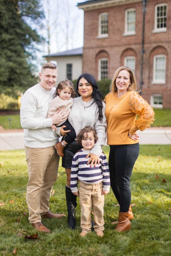 Five people pose for a family portrait on a grassy area in front of the Belair Mansion. Two adults on the left hold young children, while another adult stands on the right.