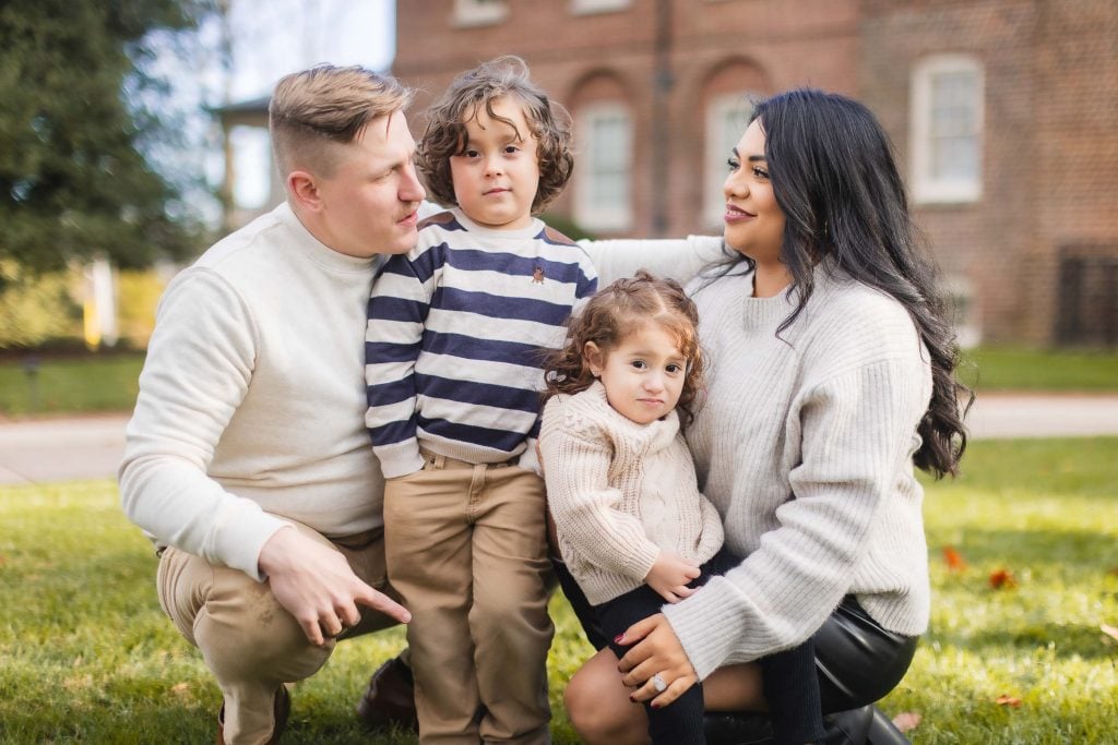 A family of four kneels on the grass in front of Belair Mansion, with two adults and two children dressed in casual autumn clothing. The woman and one child look at the camera while the man and the other child look elsewhere, capturing a charming family portrait.