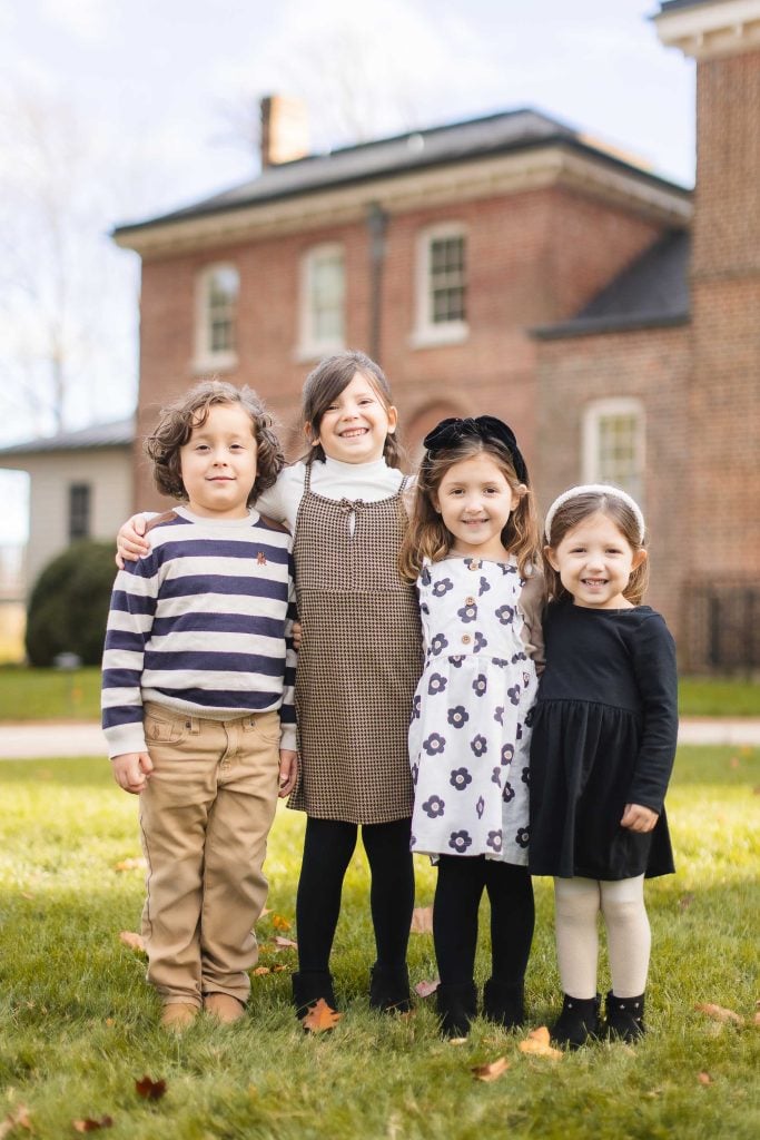 Four children stand side by side on the grass in front of Belair Mansion, smiling at the camera. The scene resembles a timeless family portrait with two girls in dresses and the other two children in longsleeve tops and pants.