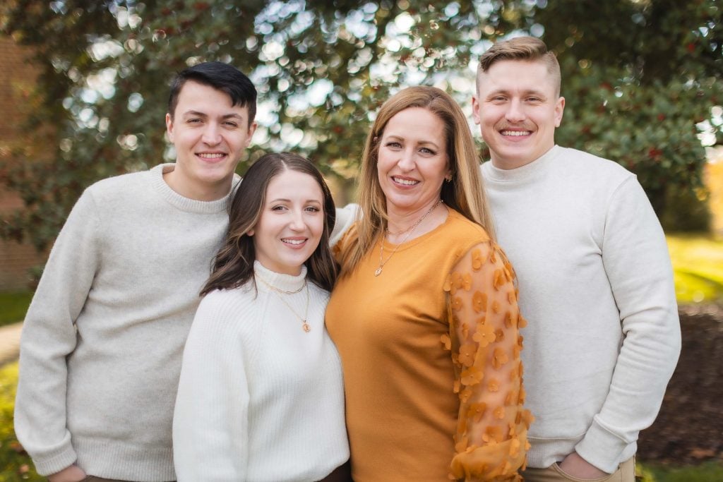 Four people, two men and two women, stand closely together smiling in front of a tree with green leaves. The women are in the center, one wearing a white sweater and the other wearing an orange blouse. This family portrait was taken at the historic Belair Mansion.