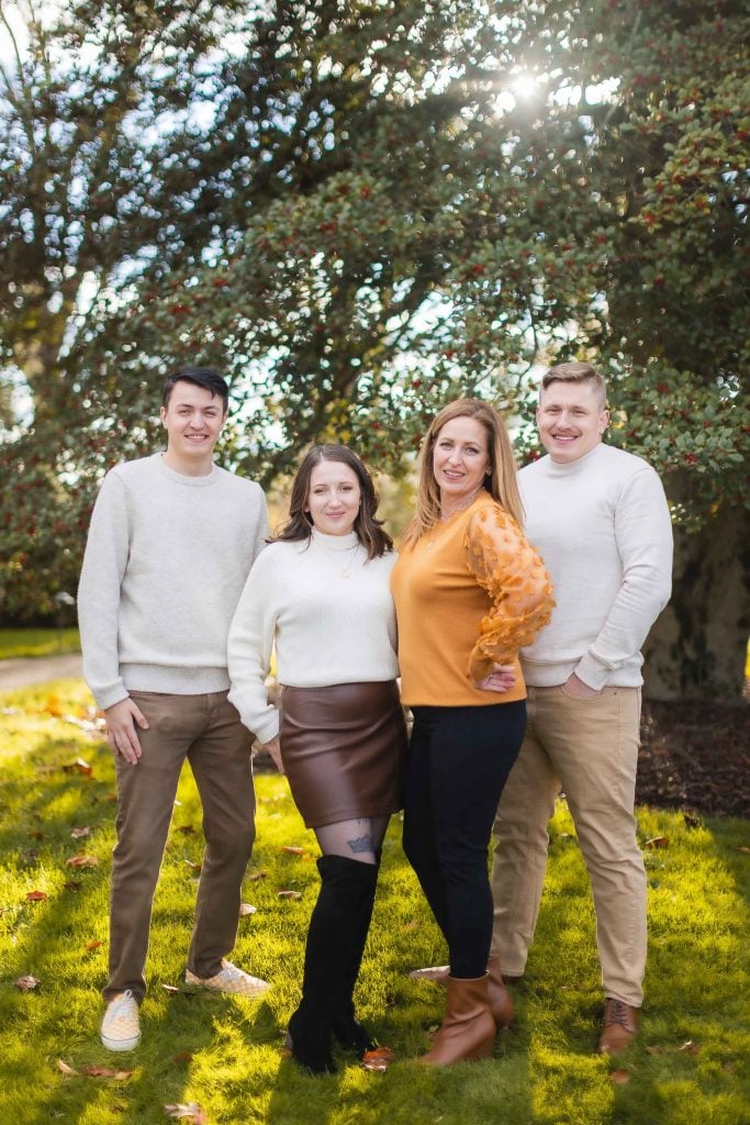 Four people stand together outdoors in casual clothing, smiling at the camera with sunlight filtering through trees in the background, creating a picturesque family portrait.