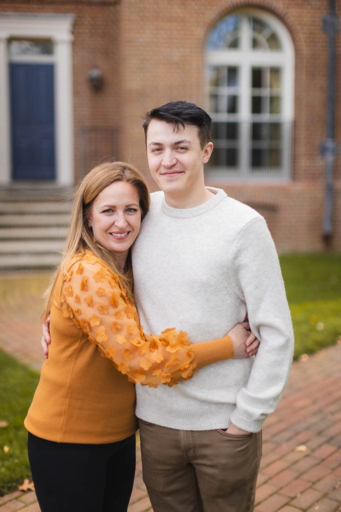 A woman in an orange sweater hugs a young man in a white sweater while standing outside the Belair Mansion, its majestic brick facade and arched windows providing the perfect backdrop for this heartfelt family portrait.