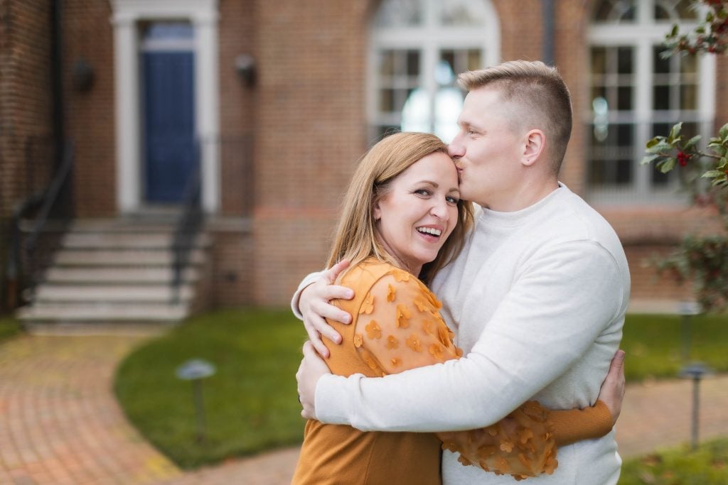 A man kisses a smiling woman on the temple while embracing her, creating a tender portrait in front of the Belair Mansion's brick facade, complete with a blue door and arched windows.