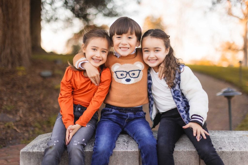 Three children sit on a stone bench outdoors, smiling at the camera in front of Belair Mansion. The child in the middle has an arm around each of the other two children. Trees and a walkway are visible in the background, creating a perfect extended family portrait setting.