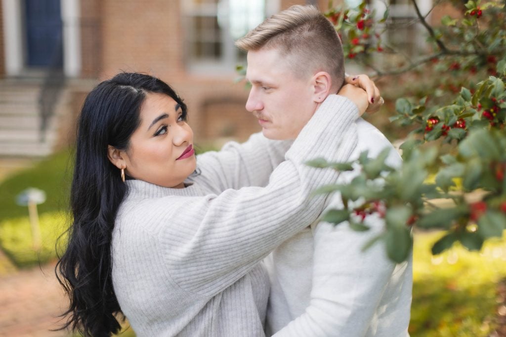 A woman and man, both wearing lightcolored sweaters, stand close together outdoors at Belair Mansion, with the woman looking at the man and holding him around his neck. Red berries and green leaves are visible nearby, creating a picturesque portrait.