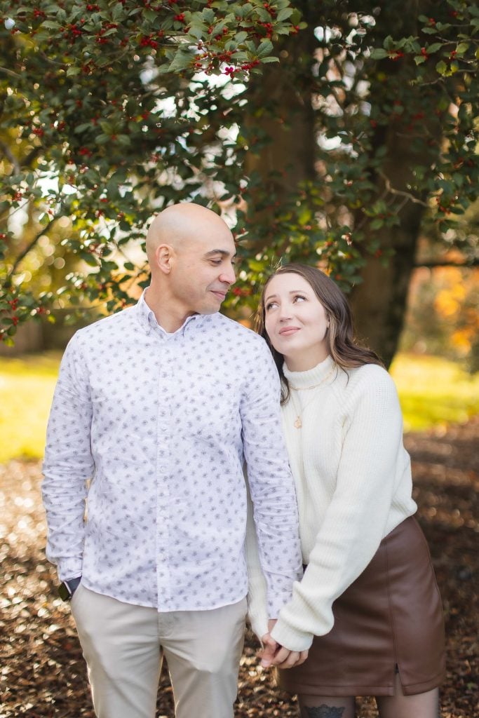 A couple stands under a tree at Belair Mansion. The woman, in a white sweater and brown skirt, looks up at the man in a patterned shirt and beige pants. They hold hands and smile, capturing the essence of family in a beautiful portrait.