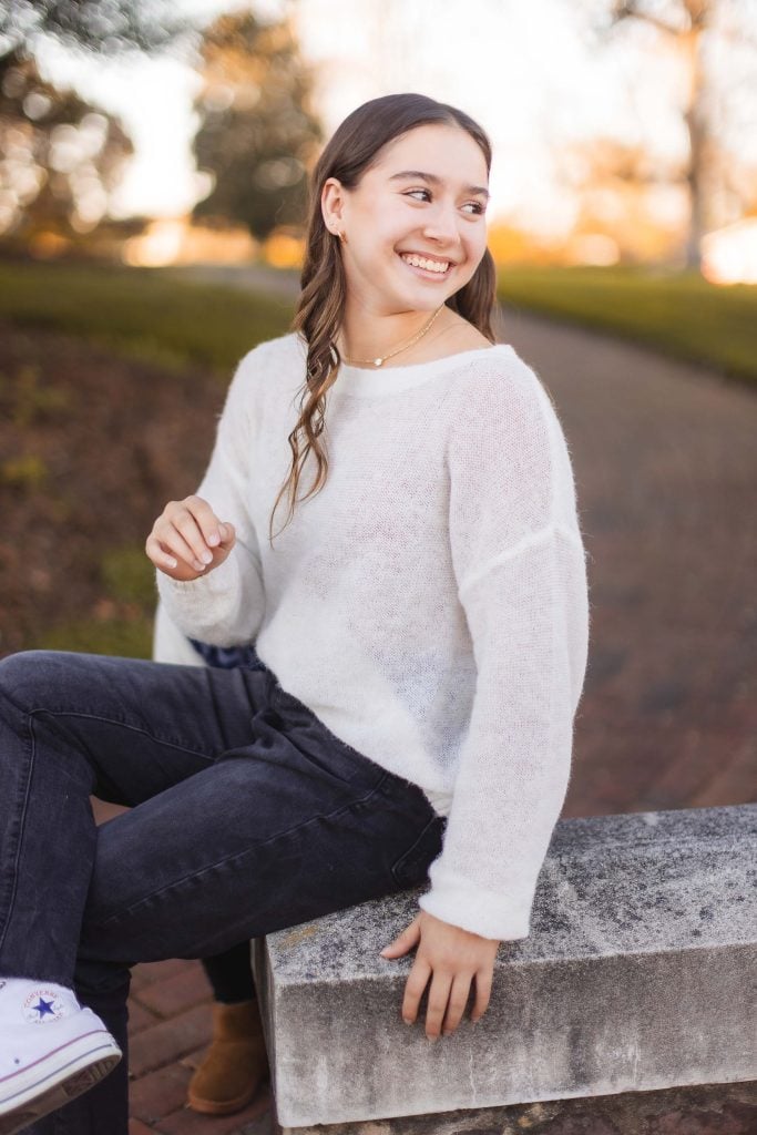 A young woman with long hair, wearing a white sweater and dark jeans, smiles while sitting on a stone bench in Belair Mansion's park.