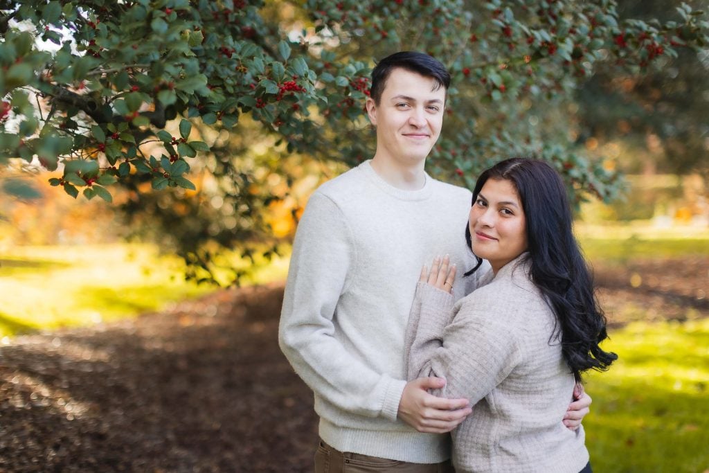 A man and a woman stand outdoors under a leafy tree, both wearing lightcolored sweaters. The woman leans against the man as they look at the camera, creating a family portrait reminiscent of serene moments one might capture at Belair Mansion.