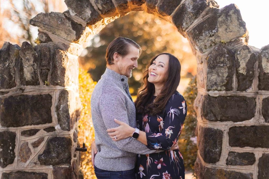 A man and woman stand in a stone archway at Belair Mansion, embracing and looking at each other happily. Trees and sunlight are in the background.