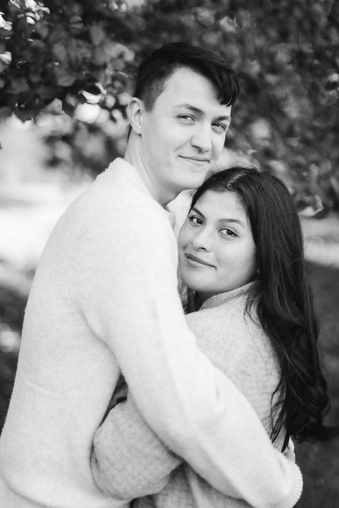 A young couple, both in lightcolored tops, embrace outdoors with greenery in the background at Belair Mansion. The woman has long dark hair, and they both look at the camera, capturing a timeless portrait.