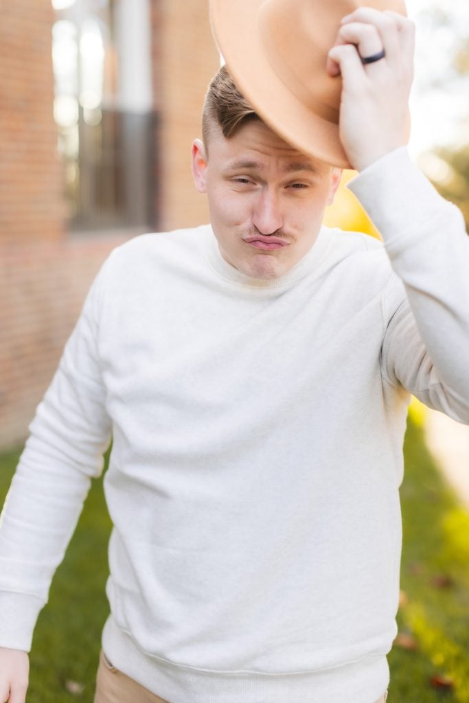 A man in a white sweater tilts his tan hat with one hand while making a playful expression, standing outside near the brick building of Belair Mansion.