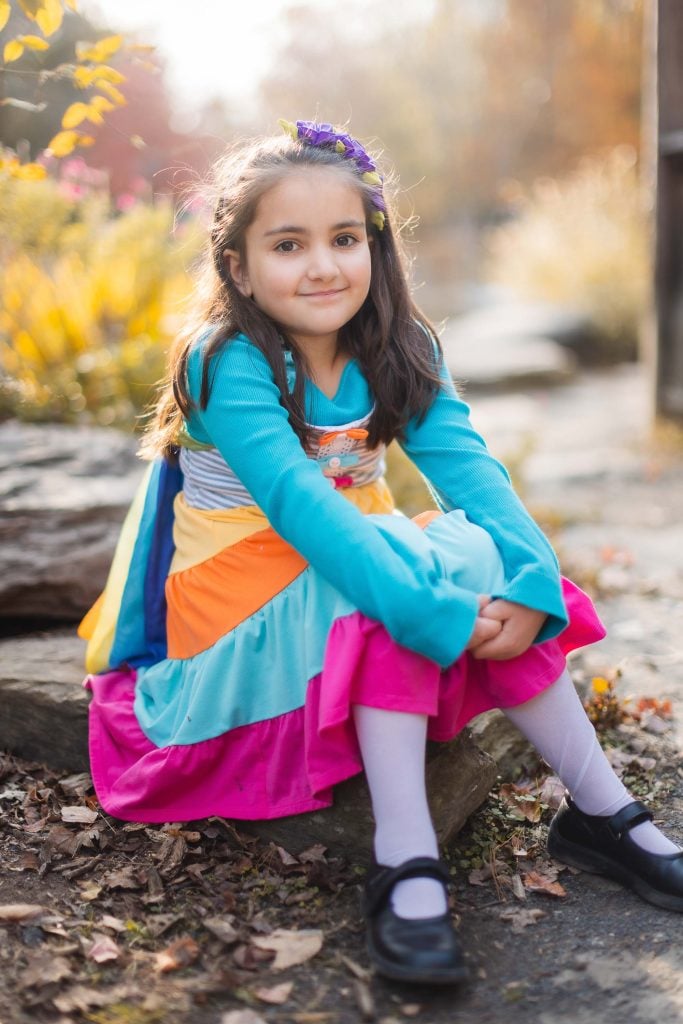 In this serene portrait, a young girl with long dark hair wearing a colorful dress and black shoes sits on a rock at Greenspring Garden, surrounded by lush trees and plants.