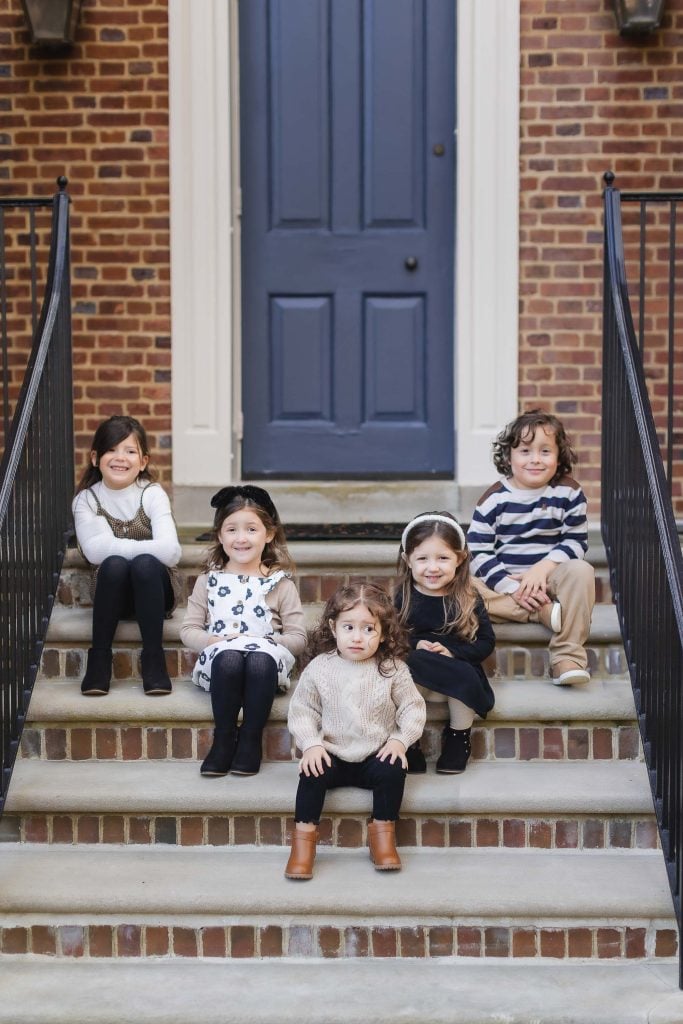 Five children sit in two rows on outdoor brick steps in front of a blue door and red brick wall, capturing a charming family portrait reminiscent of times spent at the historic Belair Mansion.